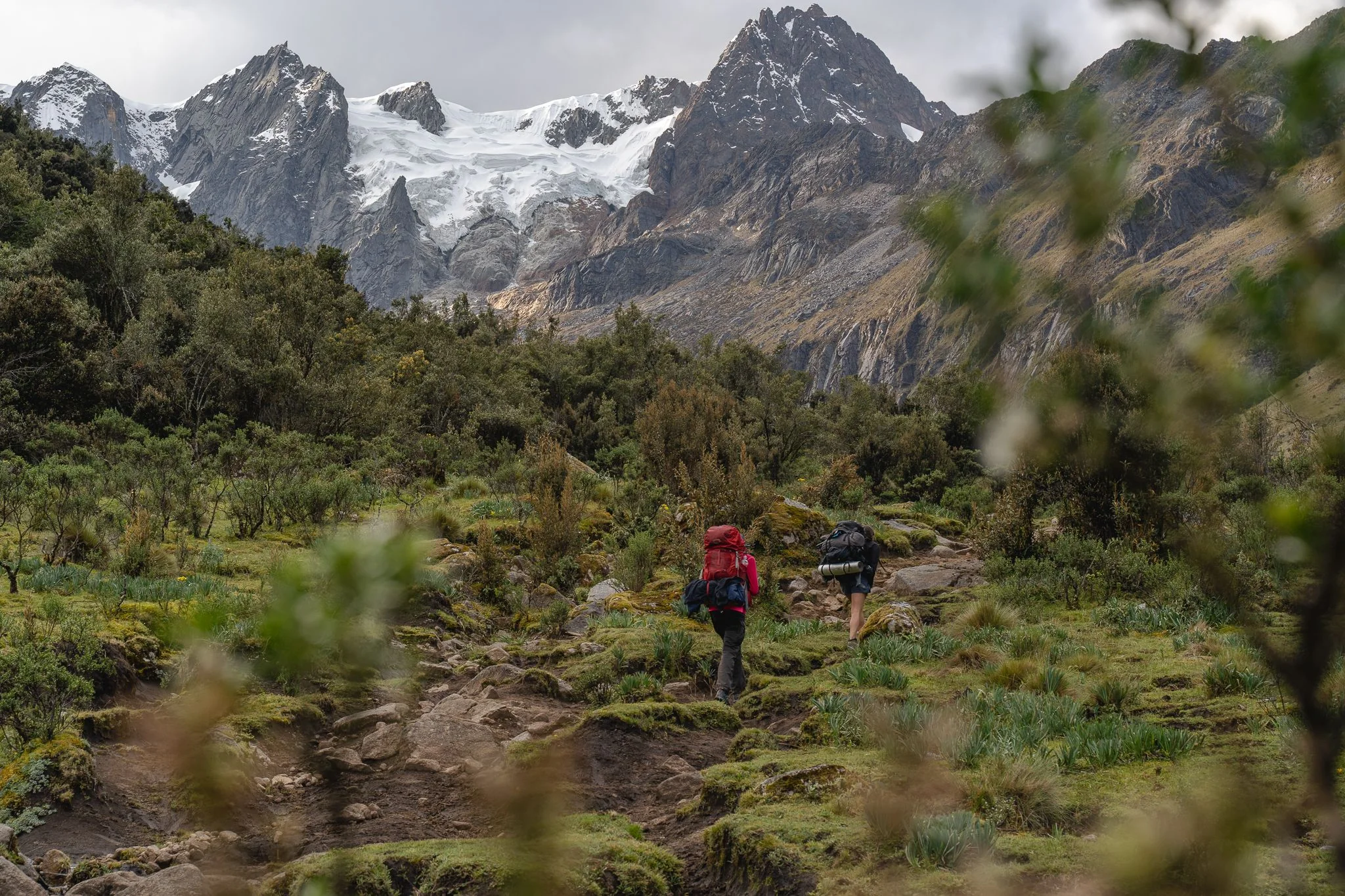 Two trekkers with large packs walking a stony trail through scrub, glaciated peaks at the head of the valley ahead, first day of the Santa Cruz trek, Peru