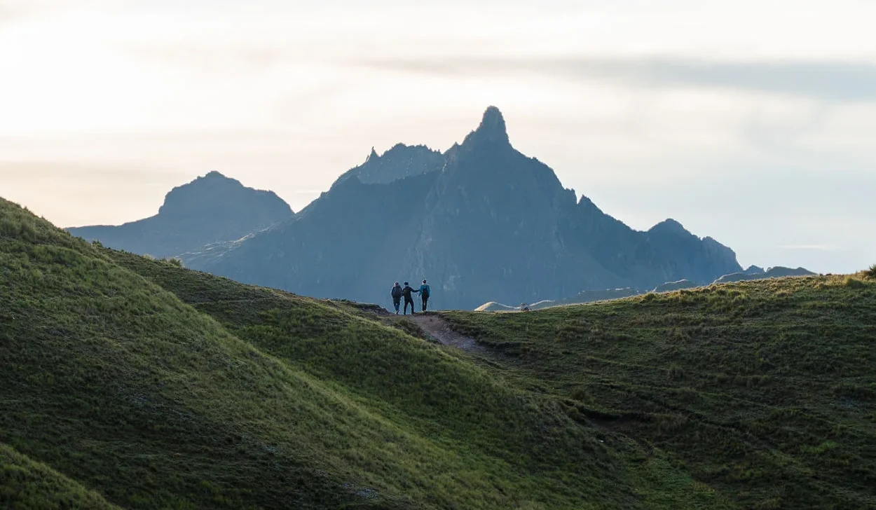 Two figures walking a green ridge with a jagged rock peak rising behind them, Ancascocha trek, Cusco region, Peru