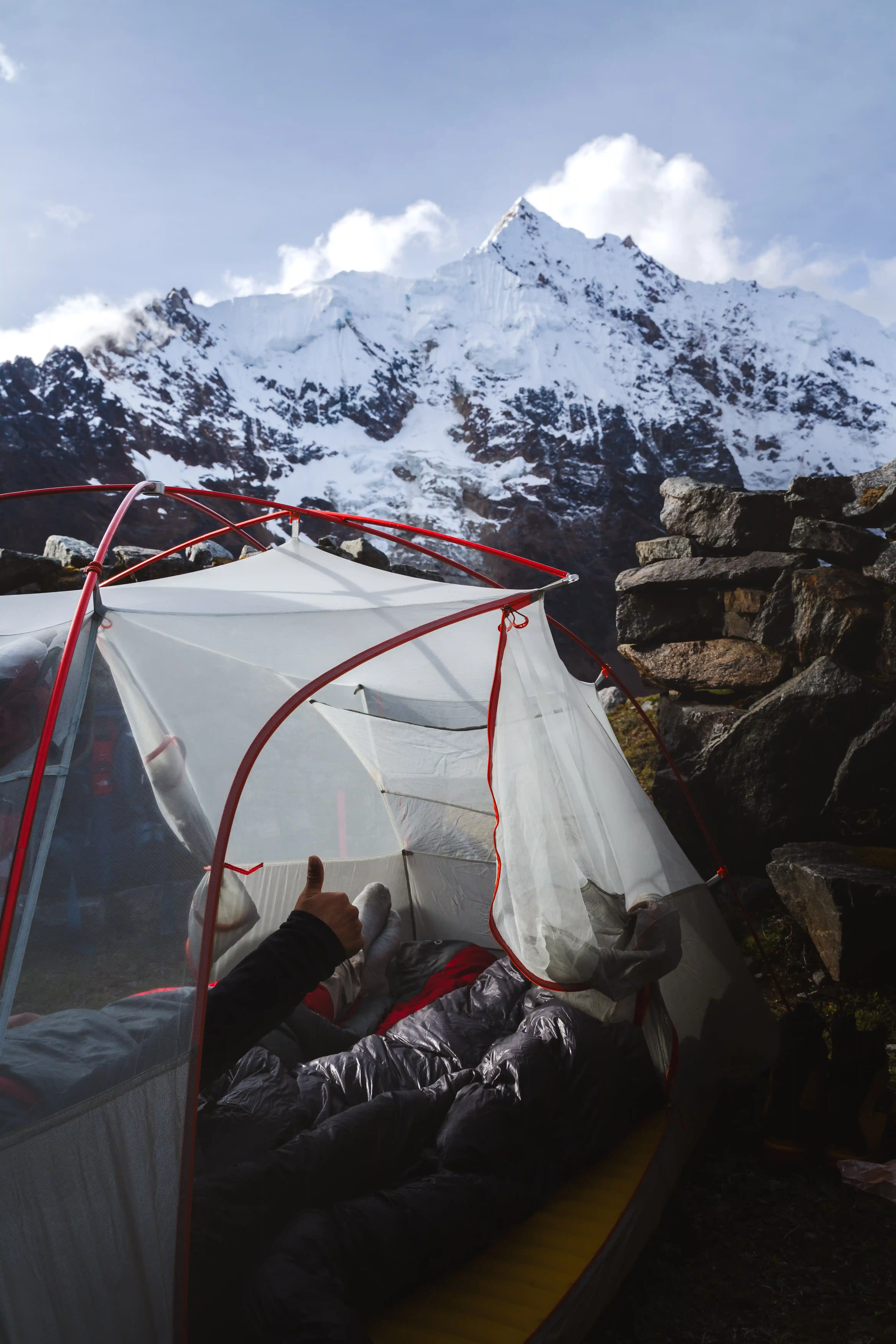 Looking out of a tent doorway from inside a sleeping bag, a climber's hands giving a thumbs up, a snow-covered peak framed beyond the stone wall of the camp, Cerro Soray, Peru