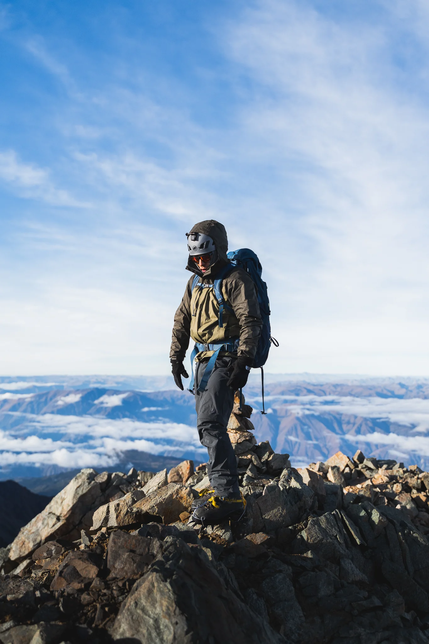 A climber in a helmet and backpack standing on a rocky summit, a sea of cloud and distant ridges below, Cordillera Vilcabamba, Peru