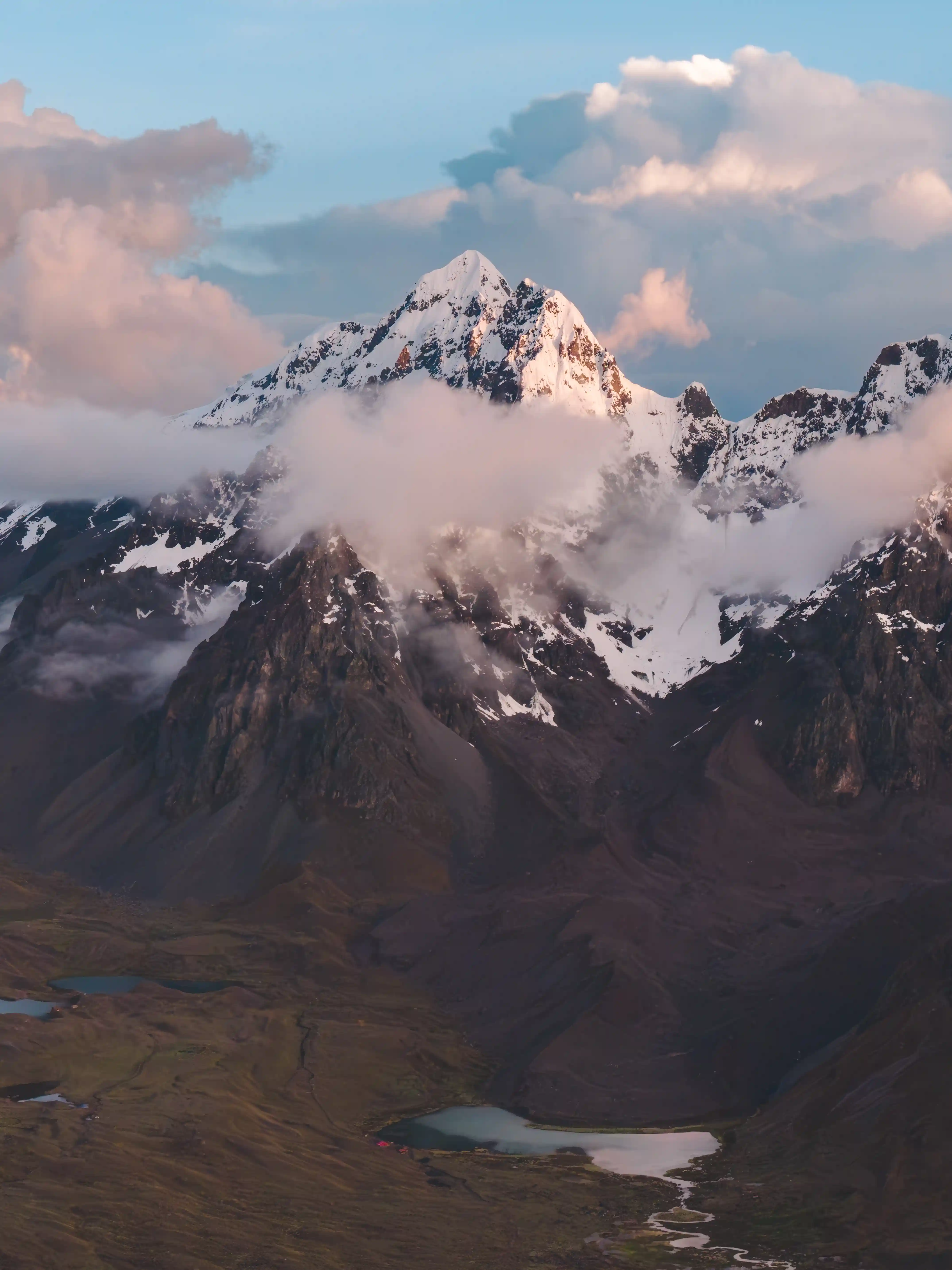 The rocky summit of Nevado Mariposa wrapped in cloud lit pink at sunset, small lakes on the dark valley floor below, Cordillera Vilcanota, Peru