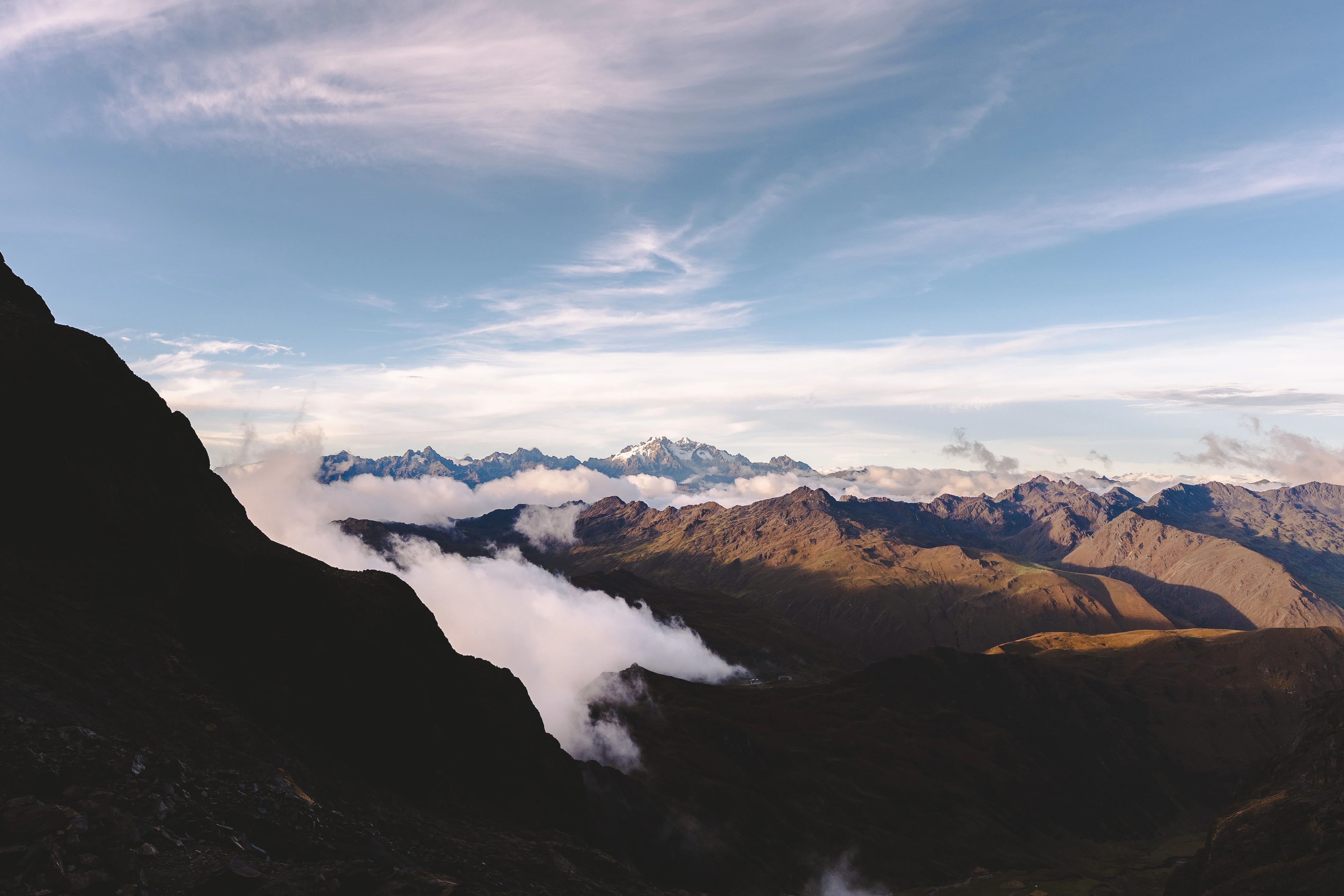 A dark mountainside in shadow above valleys filling with cloud, sunlit brown ridges and a snowy range on the horizon, Cordillera Urubamba, Peru