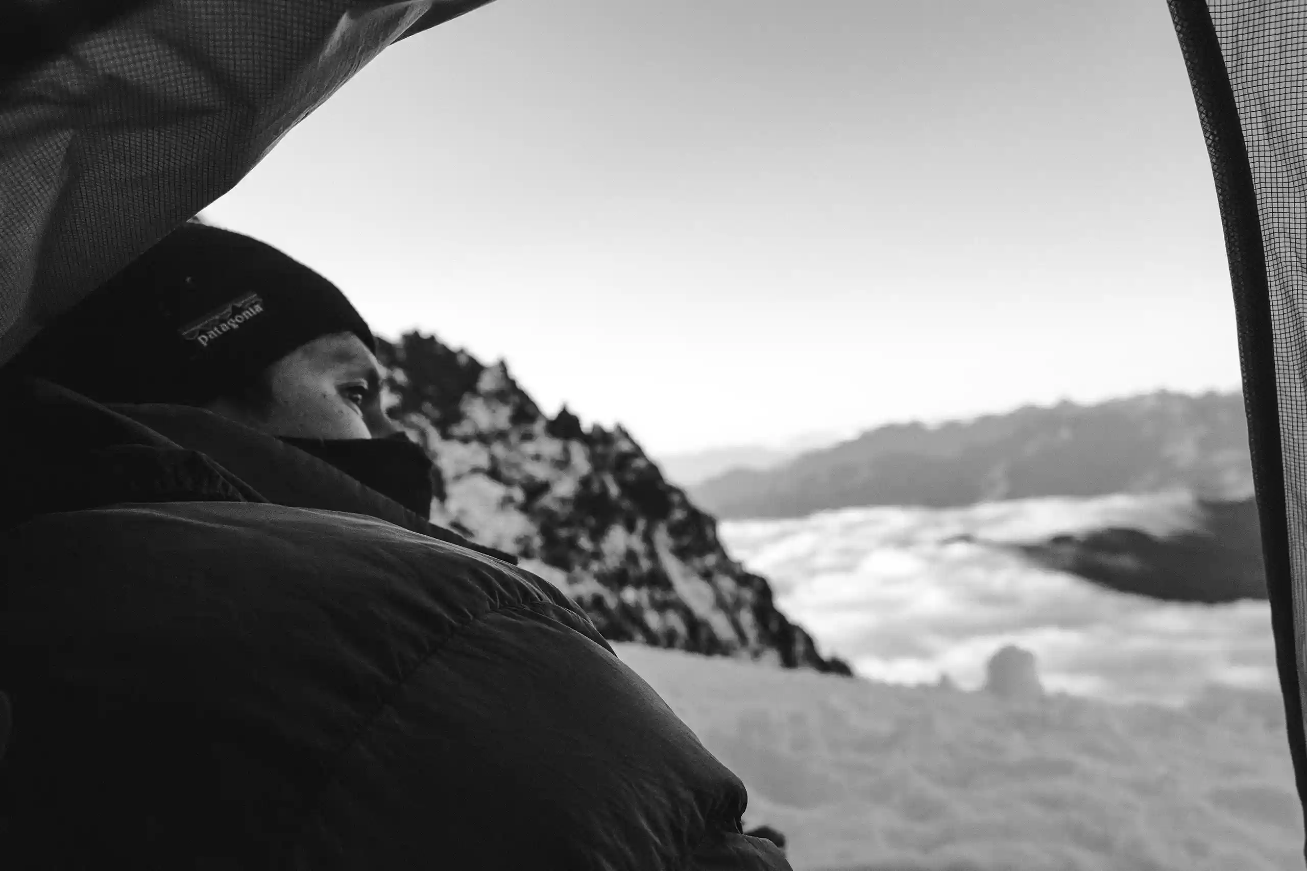Black-and-white photo of a climber in a sleeping bag looking out of a tent at snow peaks above a sea of cloud, Cordillera Urubamba, Peru