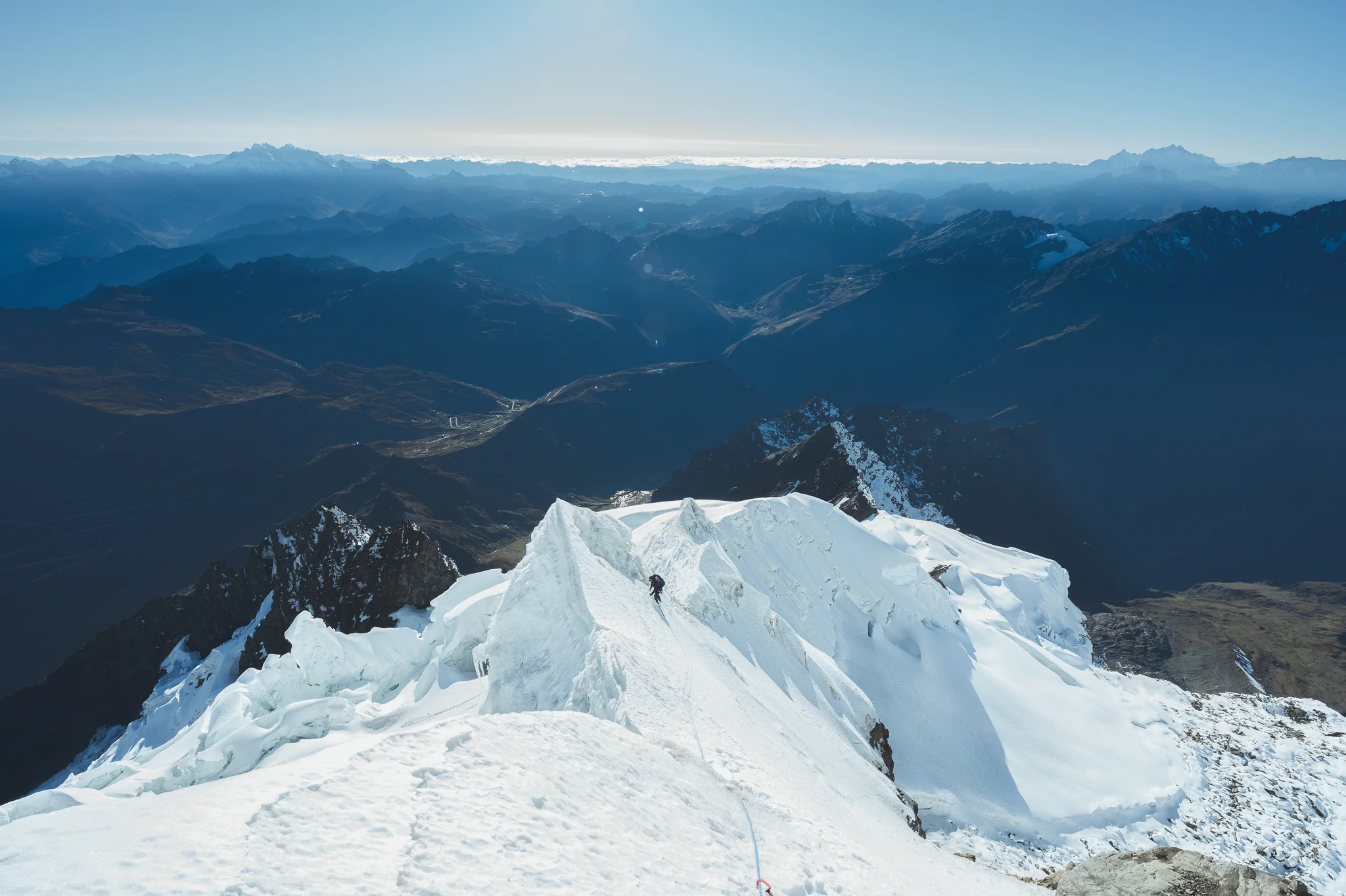A climber on a sharp, corniced snow ridge high above shadowed blue valleys and distant ranges, Cordillera Urubamba, Peru